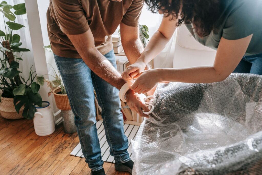 A couple wraps furniture with bubble wrap in a sunlit living room, preparing to move.