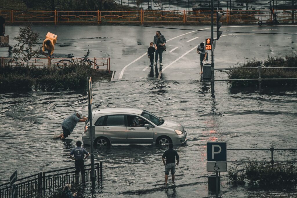 A flooded city street with a car being pushed and pedestrians navigating the water.